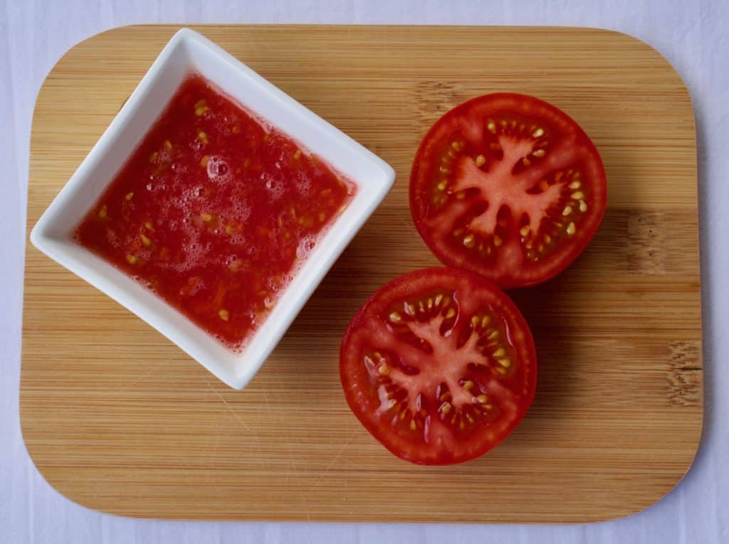 Grated tomato in a square white bowl, with two halves of a tomato next to it on a wooden board