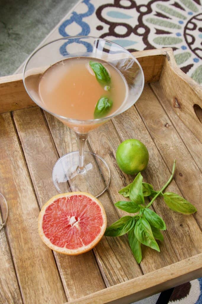 A grapefruit basil-tini on a wooden platter with a grapefruit, lime, and some basil