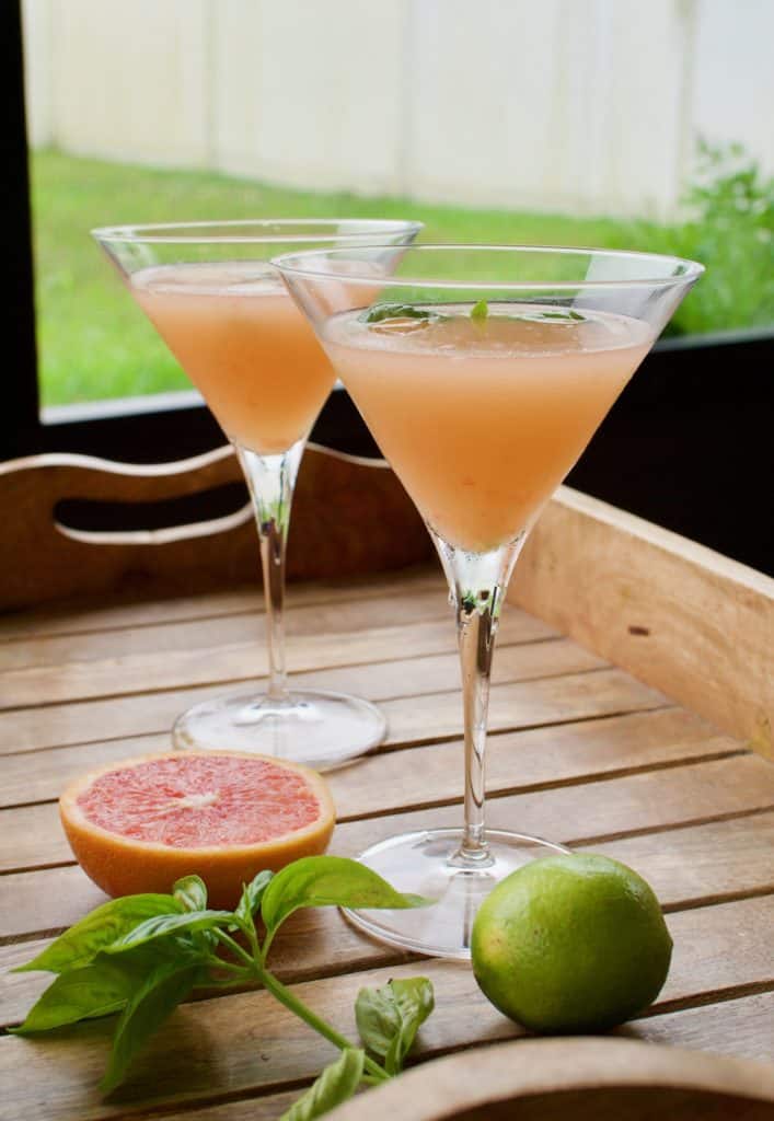 Two grapefruit basil martinis on a wooden platter with a lime, grapefruit, and some basil.