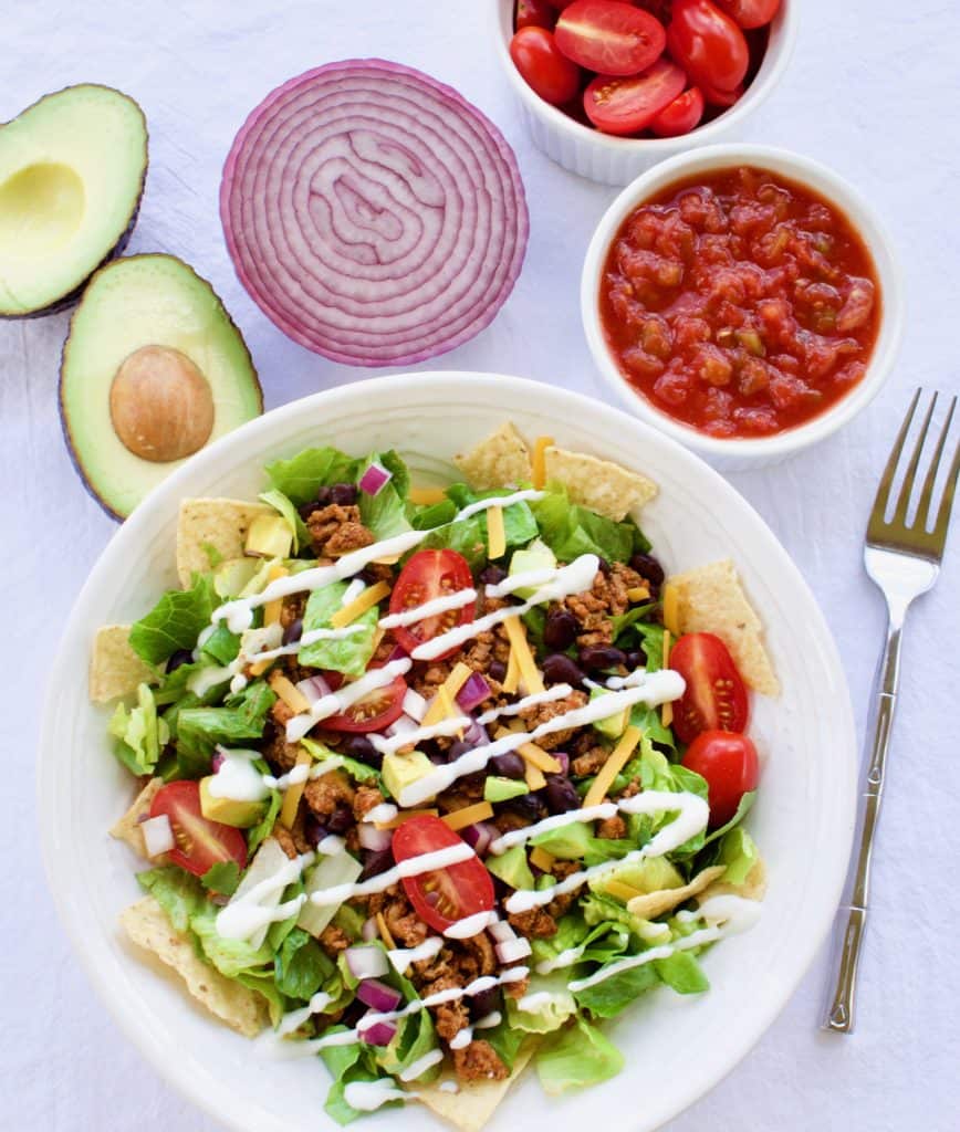Taco salad in a white bowl surrounded by ingredients, viewed from above