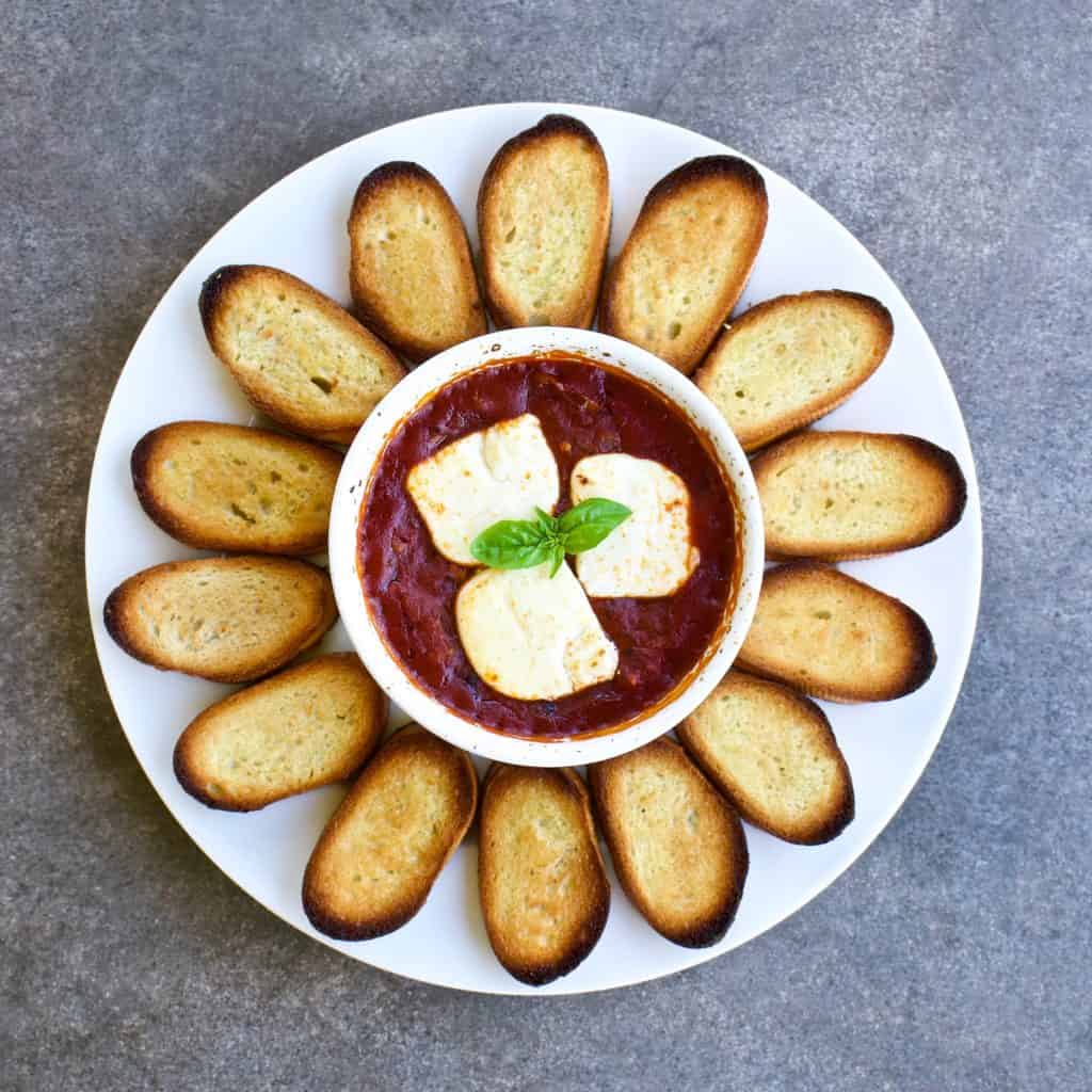 Goat cheese baked in tomato sauce served with crostini on a white plate