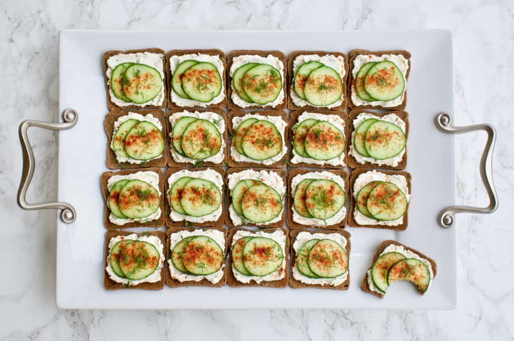 Mini cucumber toasts arranged on a serving platter. One has a bite taken out of it.