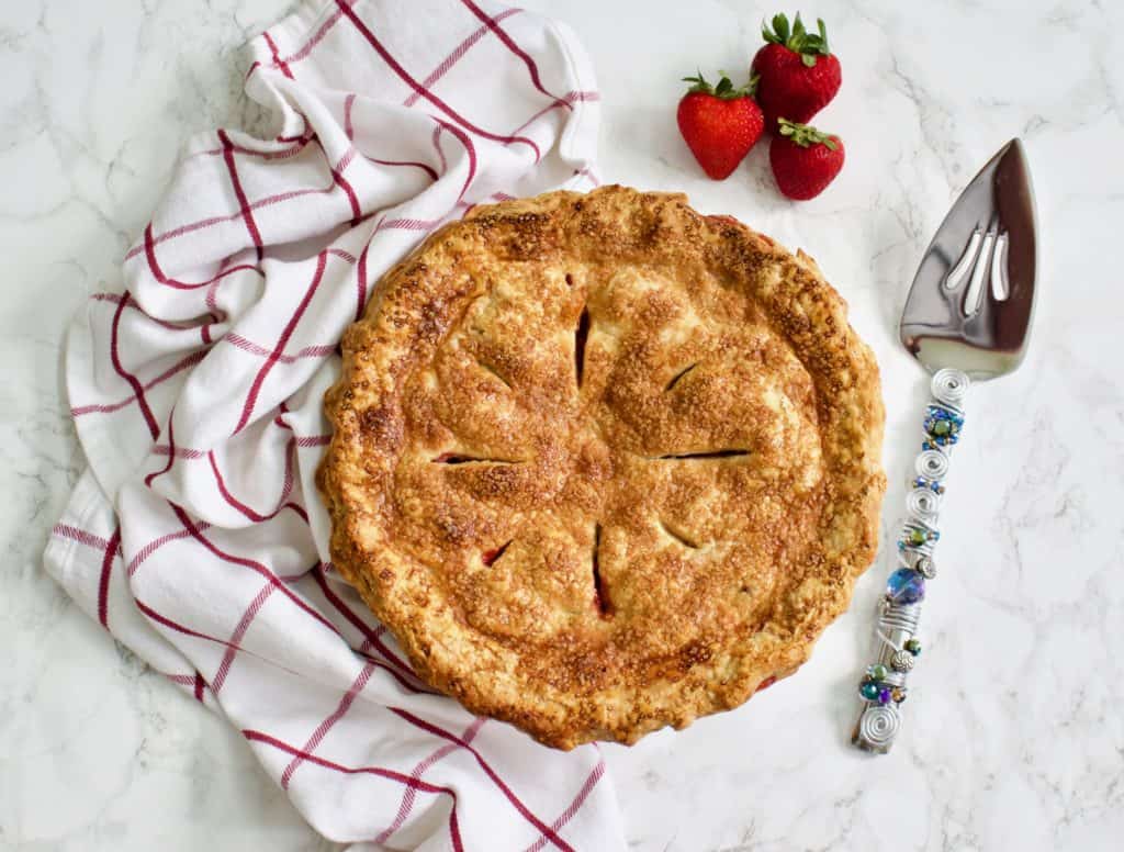 Strawberry Rhubarb Pie with red and white checkered towel, fresh strawberries and a pie server on marble surface