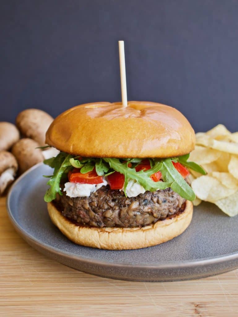 Blended mushroom and beef burger with goat cheese, roasted red peppers and arugula on a brioche bun on a gray plate with mushrooms and chips in background
